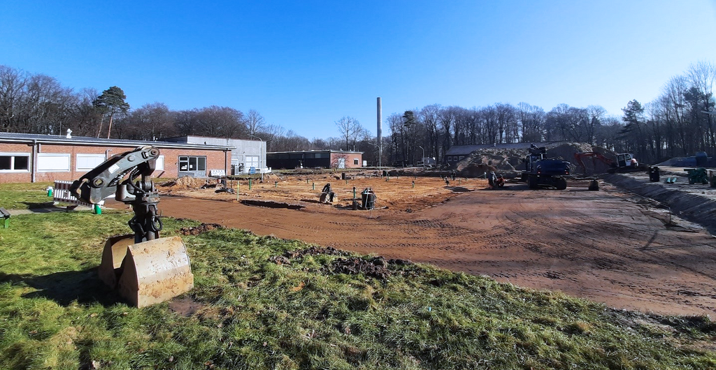Baustelle für den Gewächshauskomplex. Man sieht eine vom Bewuchs befreite, geebnete Bodenfläche und verschieden Baumaschinen.
