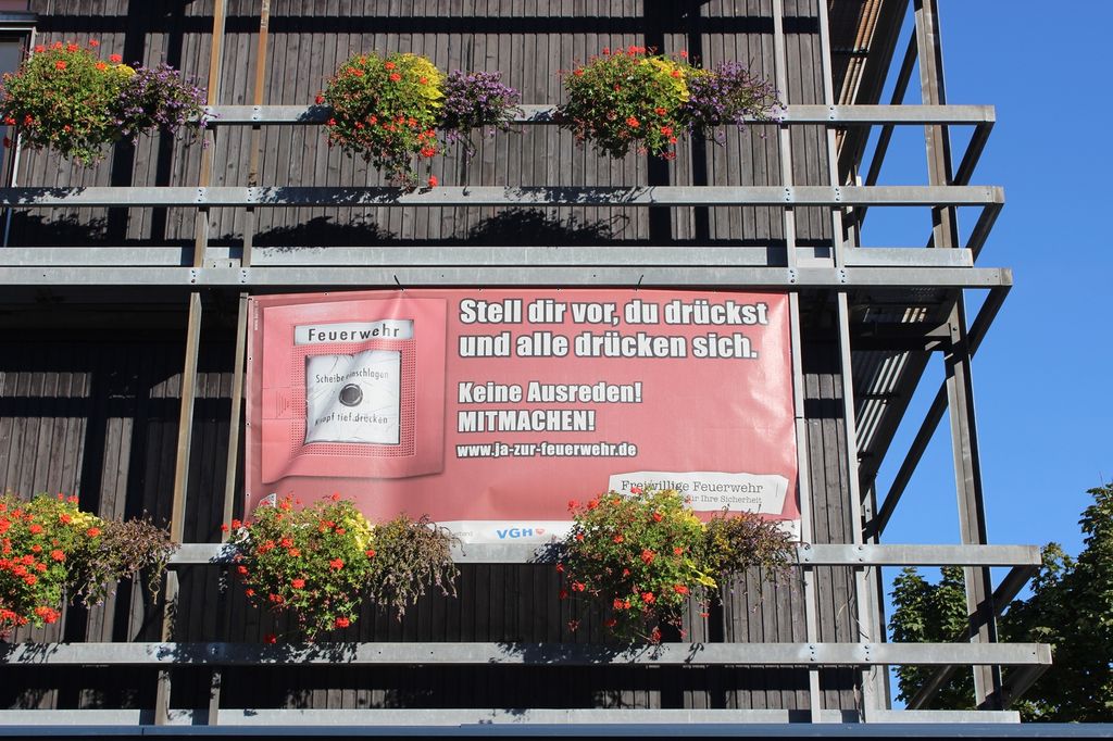 Werbeplakat des Deutschen Feuerwehrverbandes. Neben der Abbildung eines Handfeuermelders ist zu lesen: „Stell dir vor, du drückst und alle drücken sich.“