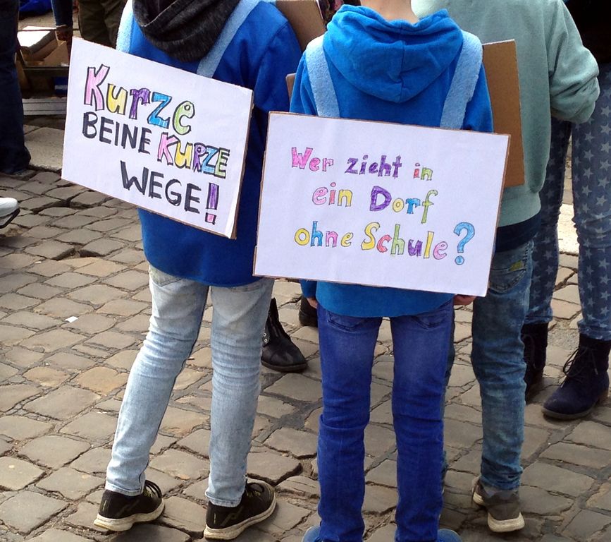 Zwei junge Menschen auf einer Kundgebung. Sie tragen Schilder mit den Aufschriften „Kurze Beine kurze Wege“ und „Wer zieht in ein Dorf ohne Schule?“